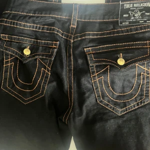 Svarta True Religion jeans Ricky Relaxed Straight - Säljer ett par svarta True Religion jeans, modell Ricky Relaxed Straight. Jeansen har kontrasterande orange sömmar, klassiska bakfickor med lock och guldiga knappar. Perfekta för dig som gillar streetwear och vill sticka ut med detaljer.