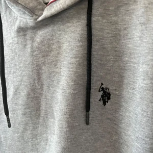 Grå hoodie från U.S. Polo Assn. - Säljer en stilren grå hoodie från U.S. Polo Assn. med svart broderad logga på bröstet. Tröjan har svarta dragsnören, magficka och ribbade muddar. Perfekt för en avslappnad look.