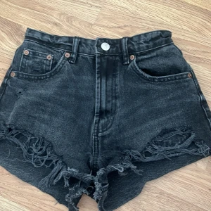Svarta jeansshorts med fransar från Zara - Snygga svarta jeansshorts från Zara med hög midja och slitna fransar nertill. Klassisk femficksmodell och knappgylf. Perfekta för en cool och avslappnad stil. I storlek 32