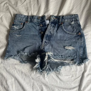 Blå jeansshorts med fransar från Zara - Säljer ett par blå jeansshorts från Zara med slitna detaljer och fransig kant. Klassisk femficksmodell med knappar framtill. Perfekta för sommaren och ger en cool, avslappnad look. Pris kan diskuteras 💖💖