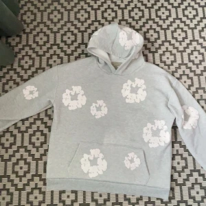 Ljusgrå hoodie med vita blommor från Denim Tears - Säljer en ljusgrå hoodie från Denim Tears med vita blommönster tryckta över hela tröjan. Tröjan har huva och en stor magficka framtill. Perfekt för dig som gillar streetwear och vill sticka ut med en unik design.