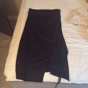 Svart omlottkjol med slits - Snygg svart omlottkjol med slits och knytband i sidan. Kjolen har en stilren design och är perfekt för dig som gillar en enkel men trendig look. Passar till många olika tillfällen och är lätt att matcha med olika toppar. Ny skick, aldrig använd, ställ frågor om de behövs 💕