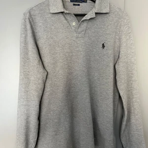Grå långärmad pikétröja från Ralph Lauren - Klassisk grå långärmad pikétröja från Ralph Lauren med krage och knappar framtill. Tröjan är i  fint skick. Storlek M, slim fit