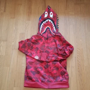 Röd camouflage zip-hoodie med hajmotiv från BAPE - Säljer en röd camouflage hoodie från BAPE med dragkedja och hajansikte på huvan. Hoodien har broderade detaljer och fickor framtill. Perfekt för dig som vill sticka ut med streetwear-stil.