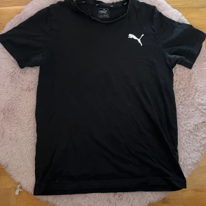 Svart t-shirt från Puma - Säljer en svart t-shirt från Puma med klassisk logga i vitt på bröstet. T-shirten har rund hals och korta ärmar, perfekt för en sportig och avslappnad stil. Tillverkad i mjuk bomull för skön komfort.