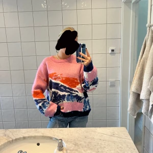 Färgglad stickad tröja med mönster - Säljer en mysig stickad tröja i rosa med mönster i blått, vitt och orange. Tröjan har rund halsringning och långa ärmar. 