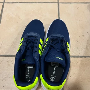 Adidas sneakers i marinblått och neon - Snygga Adidas sneakers i marinblått med neon-gula detaljer och klassiska tre ränder på sidorna. Skorna har snörning och vit platt sula. Perfekta för träning eller vardagsstil.