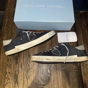 Philippe Model blå sneakers med vita detaljer - Säljer ett par Philippe Model sneakers i blå mocka med vita detaljer och snörning. Skorna har en vit platt sula och rund tå. Klassisk design med märkets logga på sidan. Perfekta för dig som gillar stilrena och bekväma sneakers.