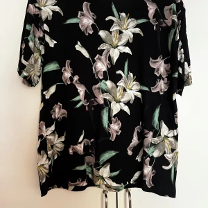 Svart blommig blus - Svart blus med stora vita och ljuslila liljor samt gröna blad. Blusen har korta ärmar och en lös passform, perfekt för en avslappnad och stilren look.