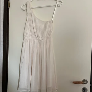 one shoulder klänning - Säljer en ljusvit kortklänning med one shoulder-design och plisserad överdel. Klänningen har ett luftigt, flowy tyg och markerad midja, perfekt för somriga tillfällen eller studentfirande.