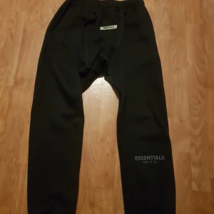 Svarta tracksuit från Essentials Fear of God med tags också  - Svarta tracksuit från Essentials Fear of God med tryckt logga på benet och litet märke framtill i midjan. Tracksuit har elastisk midja och är tillverkade i mjukt material för en avslappnad look.