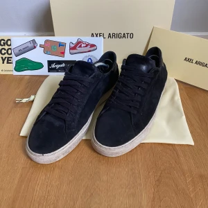 Axel Arigato skor - Snygga svarta sneakers från Axel Arigato med ovandel i mocka och klassisk vit platt sula. Skorna har rund tå och snörning framtill. Perfekta för en stilren och modern look.