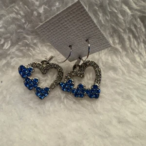 Hjärtformade örhängen med blå och silverfärgade stenar - Säljer ett par hjärtformade örhängen dekorerade med glittrande blå och silverfärgade stenar. Perfekta för att ge din look lite extra färg och glans. Hängande modell med öppen hjärtform och små blå hjärtan längs kanten. Krokarna är i sterling silver 