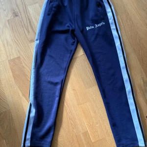 Palm Angels byxor - Säljer ett par blå byxor från Palm Angels med vita sidoränder och logga på vänster ben. Byxorna har resår i midjan och dragkedjor vid bensluten. Perfekta för en sportig och trendig look. Skick 9/10 då den är lite noprig 😀 
