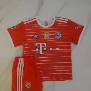 Bayern München fotbollströja med shorts - Röd och vit randig Bayern München fotbollströja med matchande shorts. Tröjan har klubbmärke, sponsorlogga och nummer 6 Kimmich på ryggen. Klassisk Adidas-design med vita detaljer och guldmärke på bröstet.