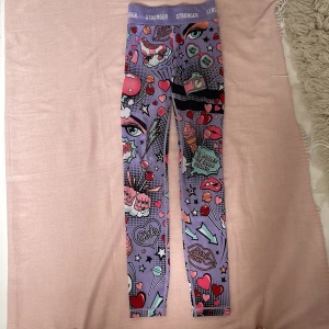 Lila leggings med pop art-mönster från Stronger - Färgglada leggings från Stronger i lila med pop art-inspirerat mönster med hjärtan, stjärnor, läppar och ögon. Hög midja och stretchigt material i återvunnen polyester och elastan. Tvättar så klart produkten innan jag skickar den. Perfekta för dig som vill sticka ut på gymmet! 