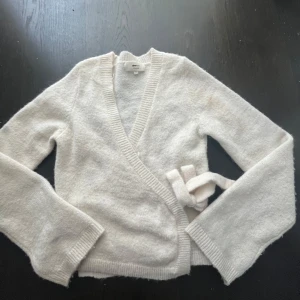 Vit omlottkofta från Gina Tricot - Mysig vit omlottkofta från Gina Tricot i ullblandning. Tröjan har långa ärmar, ribbade kanter och knytdetalj i midjan. Perfekt för lager-på-lager och en stilren look.
