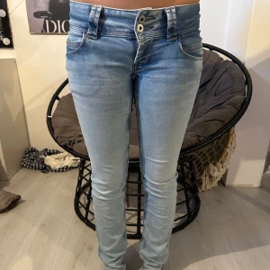 Lågmidjade pepe jeans  - Säljer ett par ljusblå jeans med dubbla knappar i midjan och klassiska fem fickor. Jeansen har en tight passform och är tillverkade i stretchigt denimtyg för en bekväm känsla.