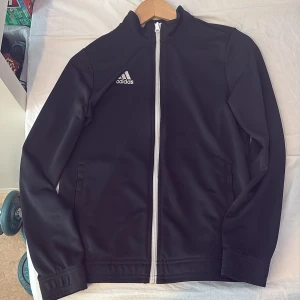 Svart träningsjacka från Adidas - Säljer en svart träningsjacka från Adidas med vit dragkedja och Adidas-logga på bröstet. Jackan har lång ärm och hög krage, perfekt för träning eller sportiga outfits. Klassisk och stilren design.