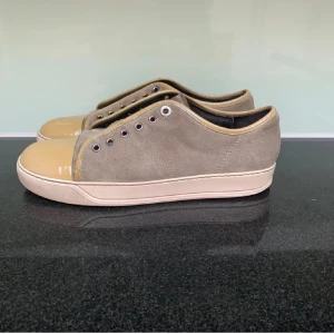 Beige och grå sneakers från Lanvin - Säljer ett par stilrena sneakers från Lanvin i grå mocka med beige detaljer och tå i blankt material. Skorna har vit sula och metallfärgade öljetter för snörning. Perfekta för dig som gillar en clean och klassisk look.