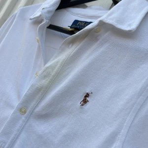 Vit skjorta från Polo Ralph Lauren - Klassisk vit skjorta från Polo Ralph Lauren med broderad logga på bröstet. Skjortan har knappar framtill och en stilren krage. Perfekt för en clean och tidlös look.