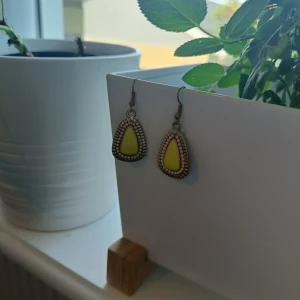 Guldfärgade örhängen med gul detalj - Säljer ett par snygga örhängen i guldfärg med droppformad gul detalj i mitten och dekorativ kant. Perfekta för att ge din outfit en färgklick och unik touch. Inte använda alls många gånger. Strass! 