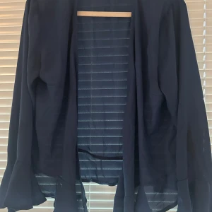 Svart cardigan jacka från Sisters Point - Säljer en svart kavaj cardigan  från Sisters Point. Kavajen/jackan har lång ärm och volangdetaljer vid ärmsluten. Den är öppen framtill och har en lös passform, perfekt att slänga över en topp.