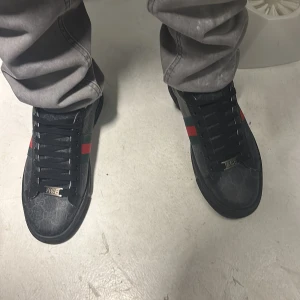 Svarta sneakers från Gucci med röd och grön rand - Säljer ett par svarta Gucci sneakers med klassisk röd och grön rand på sidan. Skorna har svarta skosnören, diskret GG-mönster i tyget och guldtexten GUCCI på hälen. Perfekta för dig som gillar lyxiga detaljer och streetstyle.