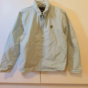 Ljusblå vindjacka från Lyle & Scott - Säljer en ljusblå vindjacka från Lyle & Scott med dragkedja och huva. Jackan har två snedställda fickor framtill och en broderad gul logotyp på bröstet. Perfekt för blåsiga dagar och enkel att matcha med olika outfits. Pris 499