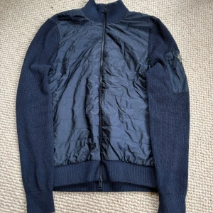 Mörkblå hybridjacka från J.Lindeberg - Snygg mörkblå hybridjacka från J.Lindeberg med quiltad front och stickade ärmar. Jackan har hel dragkedja, ribbade muddar och en diskret logga på ärmen. Perfekt för lager-på-lager och stilren look. Nypris runt 3000