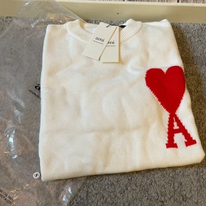 Vit stickad tröja från Ami Paris - Vit stickad tröja från Ami Paris med röd hjärtlogga och bokstaven A på bröstet. Klassisk rund halsringning och mjuk känsla. Perfekt för dig som gillar stilrena plagg med en twist. Snygg att matcha med jeans eller kjol.