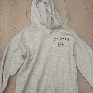 Grå hoodie Ma Chérie Gina Tricot - Supermjuk ljusgrå hoodie från Gina Tricot med svart tryck 'MA CHÉRIE' och en krona både fram och bak. Klassisk huva och långa ärmar, perfekt för chill dagar. Materialet är bomullsmix som känns skönt mot huden. Säljer då av att jag inte använder.