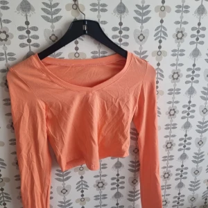 Persikofärgad croppad långärmad topp - Snygg croppad topp i persikofärgad bomull med v-ringning och långa ärmar. Perfekt för dig som gillar en enkel men trendig stil. Passar till både jeans och kjol för en chill look.