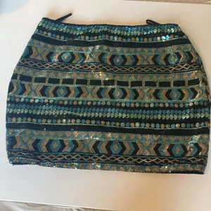 Glittrig kortkjol med paljetter - Snygg kortkjol med glittriga paljetter i blått, guld och svart. Kjolen har ett geometriskt mönster och är täckt av skimrande detaljer som verkligen sticker ut. Perfekt för dig som vill ha något extra till din outfit.