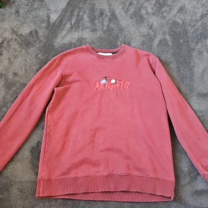 Röd sweatshirt Axel Arigato broderi - Säljer en röd sweatshirt från Axel Arigato med broderad text och blommor på bröstet. Tröjan har rund hals, långa ärmar och är gjord i mjuk bomull. Perfekt för dig som gillar streetstyle och snygga detaljer.