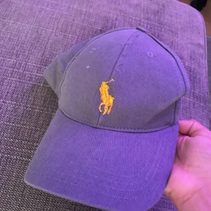 Mörkblå keps från Polo Ralph Lauren - Snygg mörkblå keps från Polo Ralph Lauren med klassisk gul broderad logga framtill och justerbart spänne bak. Kepsen är tillverkad i bomull och har en stilren, tidlös design som passar perfekt till streetwear eller casual outfits.