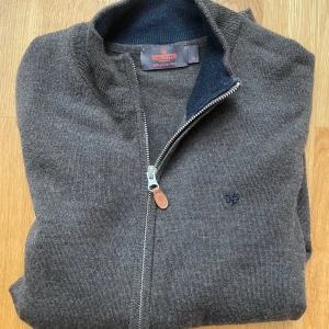 Mörkgrå merinoullskofta från Morris - Snygg brun kofta från Morris i 100% merinoull. Modellen har hel dragkedja med läderdetalj, ribbade muddar och diskret broderad logga på bröstet. Perfekt för dig som gillar stilrena och klassiska plagg med en modern touch.