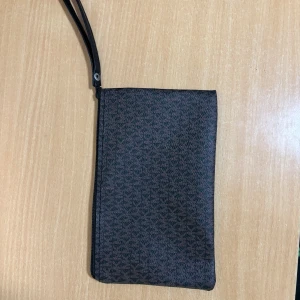 Svart kuvertväska med mönster - Stilren svart kuvertväska med diskret geometriskt mönster och handledsrem. Väskan har dragkedja upptill och är perfekt för att bära det viktigaste. Materialet känns som syntet och ytan är lätt att torka av. Enkel att matcha till olika outfits.