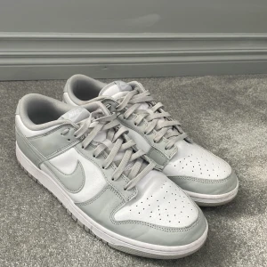 Nike Dunk Low i vitt och ljusgrått - (STORLEK 47.5) Nike Dunk Low sneakers i vitt och ljusgrått skinn. Klassisk siluett med perforerad tå, platt sula och snörning. Swoosh-logga på sidorna och broderad Nike-text på hälen. Perfekta för dig som gillar clean och stilren streetwear. Lite smutsiga men det kan man tvätt bort lätt. Använda två gånger