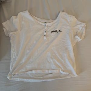 Vit croppad topp från Hollister - Säljer en vit croppad topp från Hollister med korta ärmar och rund halsringning. Toppen har knappdetaljer framtill och broderad Hollister-logga på bröstet. Skön bomullskvalitet och avslappnad passform. Tröjan är köpt second hand:) 