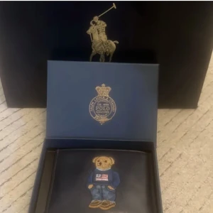 Svart plånbok Polo Ralph Lauren björn - Svart plånbok i skinn från Polo Ralph Lauren med broderad björn på framsidan som har jeans och blå tröja med amerikansk flagga. Flera kortfack och diskret Polo-logo på baksidan. Kommer i originalförpackning.