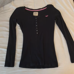 Svart ribbad långärmad tröja Hollister - Mörk blå långärmad tröja från Hollister, vintage i ribbad bomull med en tight passform. Tröjan har knappar framtill och en rosa Hollister-logga på bröstet samt rosa kant på insidan av halsringningen. Har använt 1 gång och den är köpt på plick:)