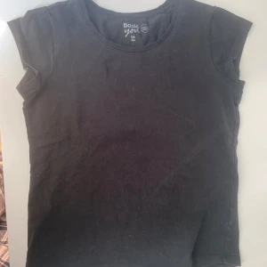 Svart basic T-shirt från Basic You - Enkel svart T-shirt från Basic You i ekologisk bomull. Modellen är rak med rund halsringning och korta ärmar, perfekt till jeans eller kjol. Stilren och lättmatchad för en clean look.