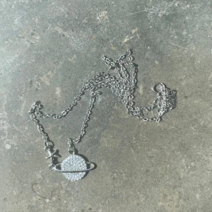Silverfärgat halsband ZEND DETAILS - Ett snyggt silverfärgat halsband med ett glittrigt planet-hänge och en liten stjärna bredvid. Kedjan är tunn och smidig, och planeten är täckt av små gnistrande stenar som ger extra bling. Perfekt för dig som gillar rymdtema och vill sticka ut lite extra.