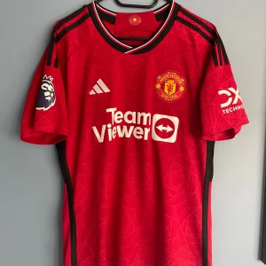 Manchester United Højlund 11 Adidas tröja - Manchester Uniteds officiella matchtröja från Adidas med Højlund och nummer 11 på ryggen. Röd med svarta detaljer, vita tryck och klubbmärke på bröstet. Premier League-logga på ärmen och TeamViewer sponsor framtill. Tillverkad i lätt och ventilerande polyester.