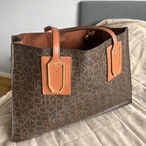 Väska från Calvin Klein - Snygg brun axelväska från Calvin Klein med monogrammönster och detaljer i ljusbrunt skinn. Väskan har rejäla handtag, guldiga metalldetaljer och kommer med en matchande innerficka. Perfekt för dig som vill ha plats för allt och ändå se stilren ut.