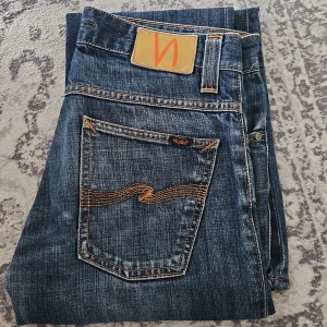 Nudie Jeans Bootcut Ola blå jeans - Säljer ett par klassiska Bootcut Ola jeans från Nudie Jeans i blå tvätt med snygga slitningar och orange kontrastsömmar. Jeansen har bootcut-passform, fem fickor och är tillverkade i 100% bomull. Jeansen kan eventuellt vara upplagda efter mig som är 170cm lång 