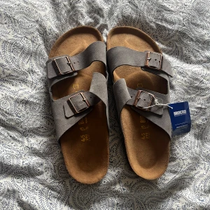 Sandaler Birkenstock - Snygga sandaler från Birkenstock i grå mocka med två breda remmar och justerbara metallspännen. Fotbädden är i mjukt skinn och sulan har korkdetaljer. Perfekta för sommardagar.