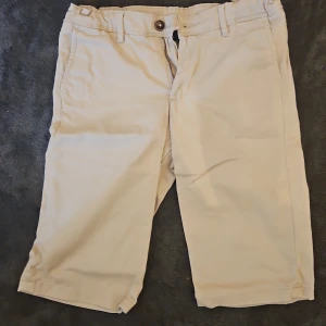 Beige shorts från Jack & Jones - Snygga beige shorts från Jack & Jones med klassisk passform och bälteshällor. De har dragkedja och knapp framtill, samt fickor både fram och bak. Perfekta för varma dagar och enkla att matcha med det mesta.