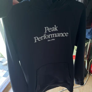 Svart hoodie från Peak Performance - Svart hoodie från Peak Performance med vit logga på bröstet. Klassisk modell med huva och känguruficka framtill. Perfekt för en chill och sportig stil. Mjuk och skön, passar till det mesta.